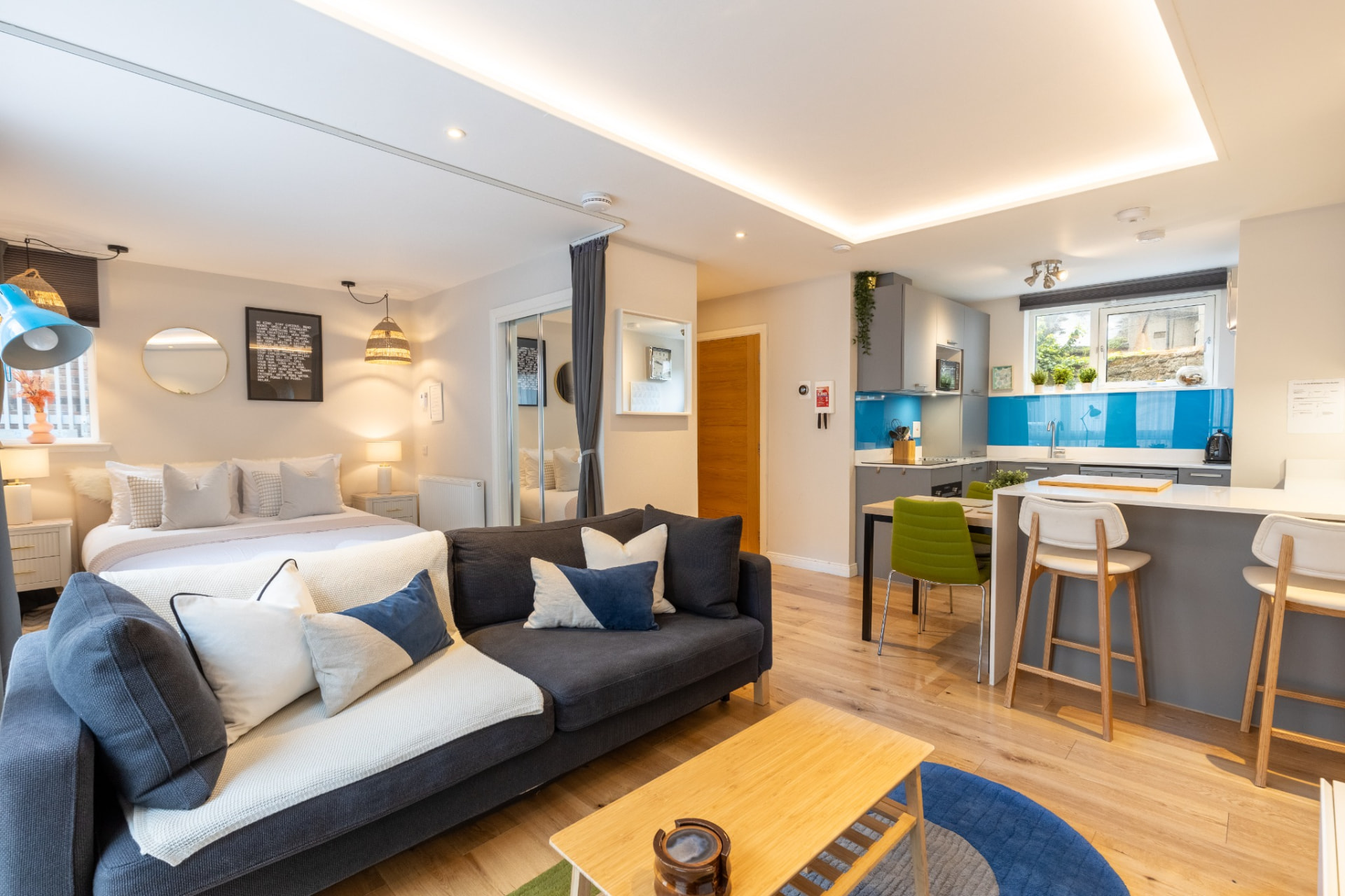 Open-plan studio at Heavenly Pad, Portobello, with a grey sofa, bed and kitchen with a blue splashback and breakfast bar.